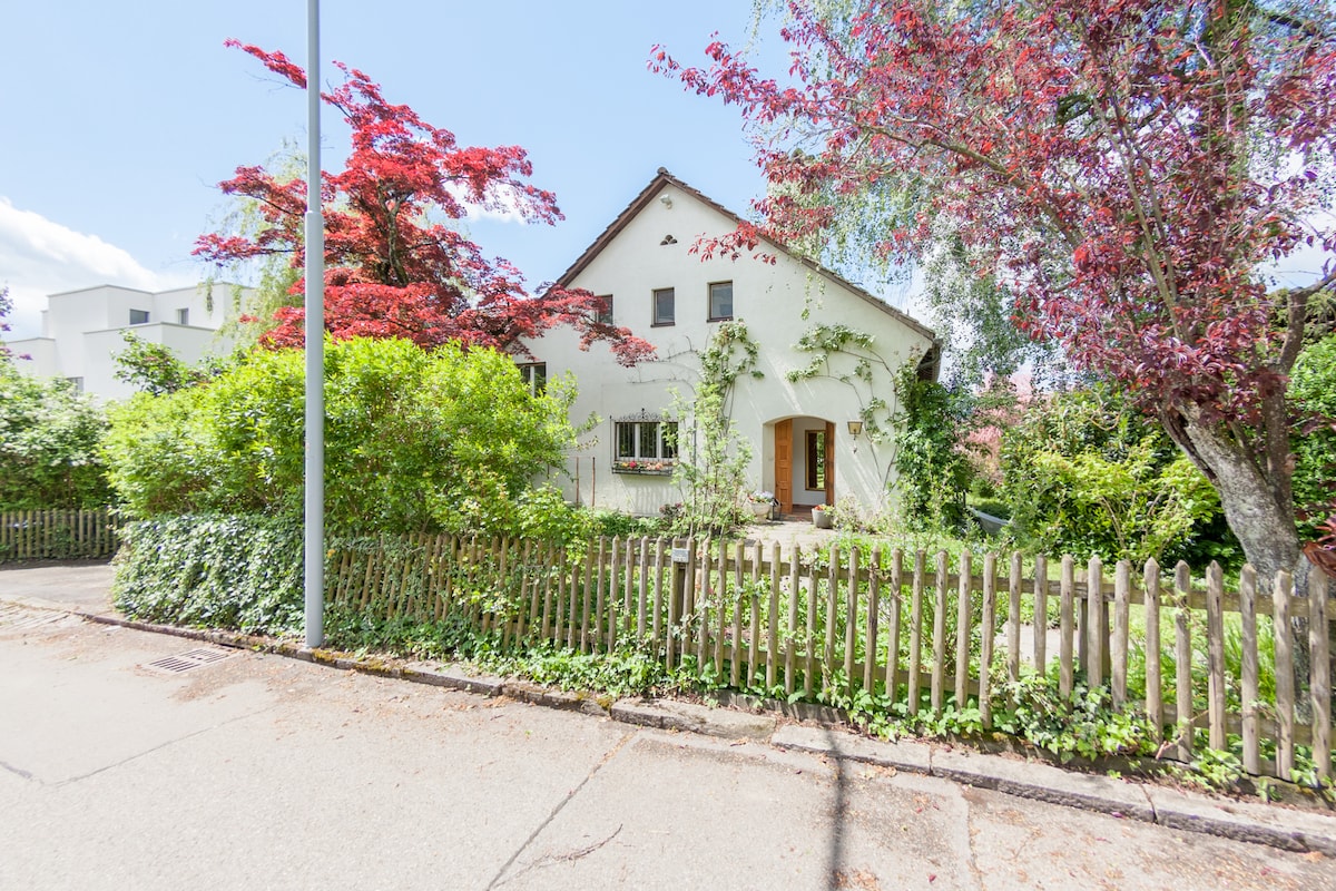 The house features a charming exterior with a light-colored facade, complemented by vibrant red foliage from nearby trees. A wooden fence surrounds the property, enhancing the inviting atmosphere. The entrance is marked by a welcoming doorway, set among lush greenery.
