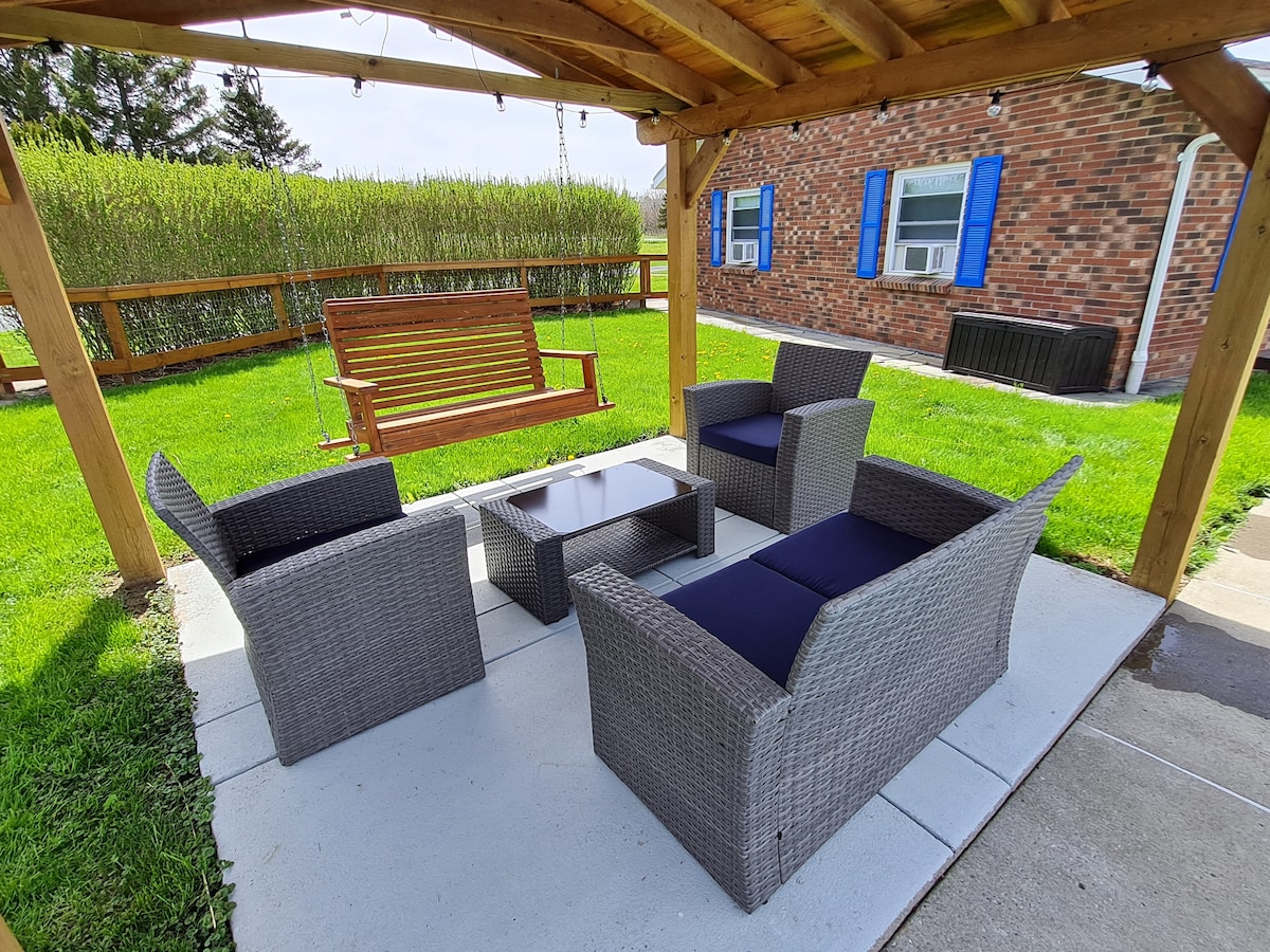 A covered outdoor seating area features four wicker chairs with dark cushions surrounding a small table, set on a concrete patio. A wooden swing is positioned nearby, with a backdrop of lush green grass and a wooden fence.