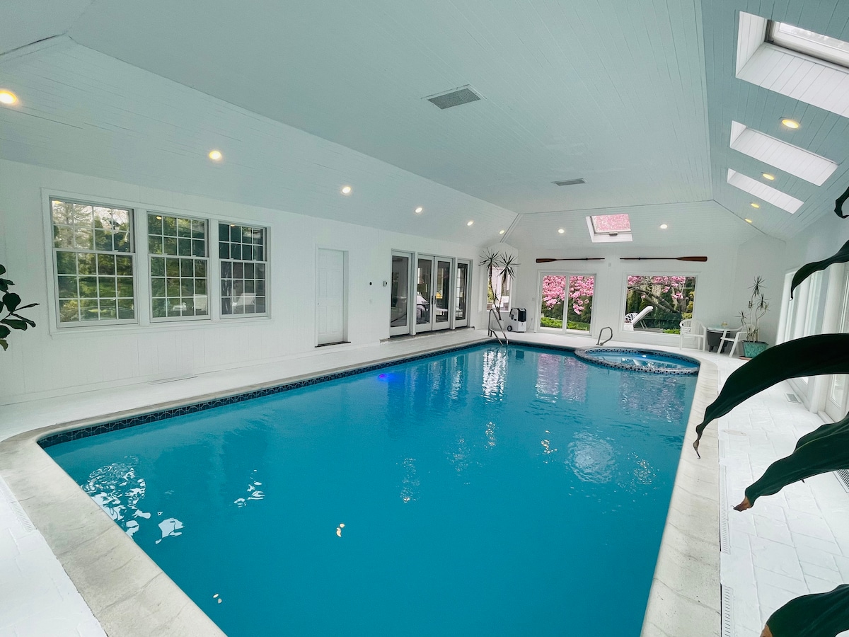 An indoor heated saltwater pool is surrounded by large windows that invite natural light. The space is accented by sleek white walls and high ceilings with skylights. A hot tub is visible near the pool, contributing to a relaxing atmosphere.