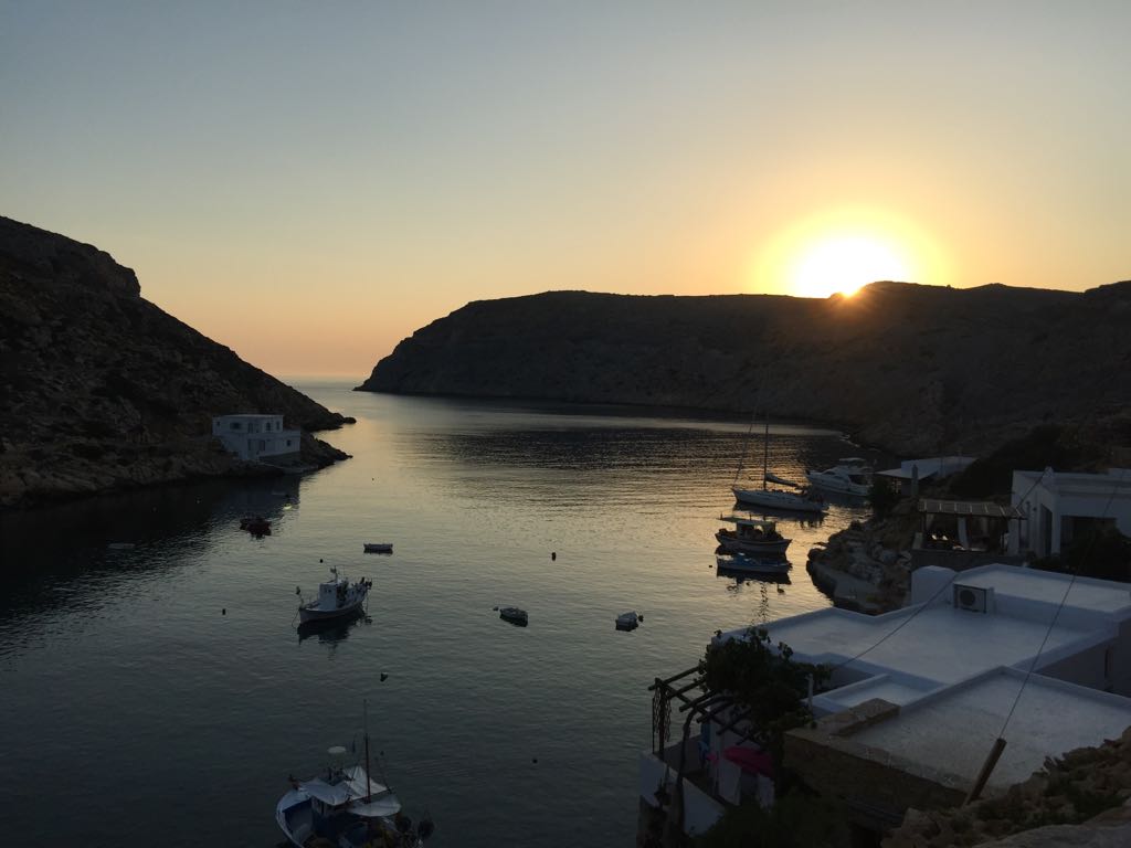 A serene coastal view is captured at sunset, with gentle waves reflecting the soft golden light. Boats are anchored in the calm waters of the bay, framed by rugged cliffs on either side. The peaceful scene invites relaxation and appreciation of nature.