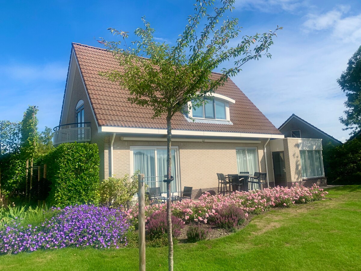 A spacious house is set in a landscaped garden, featuring vibrant flower beds and a grassy area. Large windows reflect natural light, while outdoor seating is visible on a patio. A tree stands in the foreground, enhancing the serene ambiance.