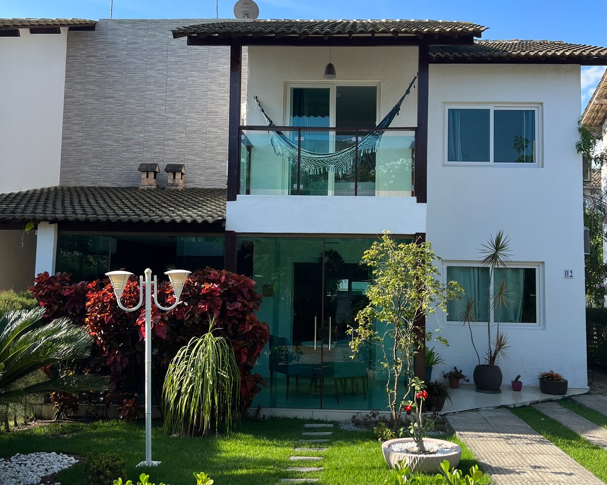 A modern bungalow-style home is depicted, featuring a two-story façade with a spacious balcony. Lush greenery surrounds the property, including colorful plants and a well-kept garden. Large glass windows enhance natural light, while a decorative lamp adds to the entryway's charm.