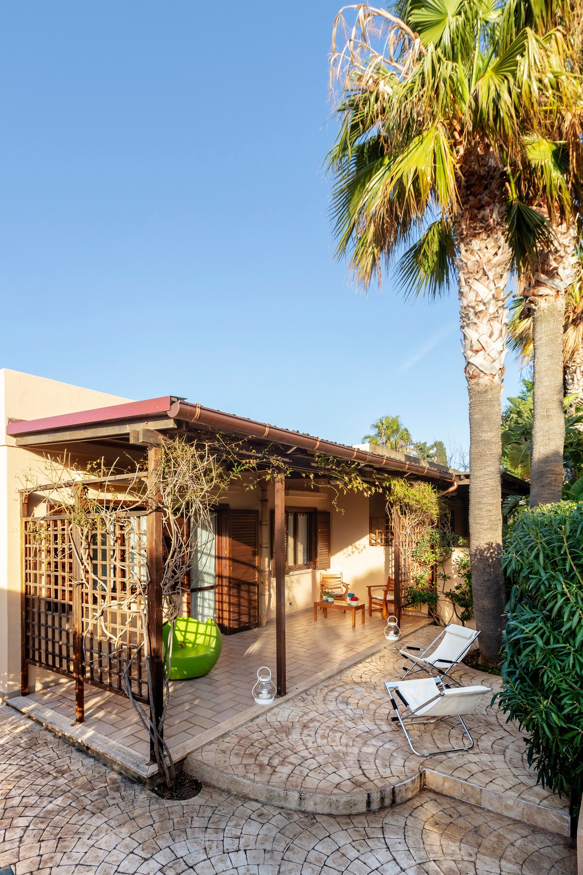A single-level villa is framed by palm trees and lush greenery. The patio features a wooden pergola with seating, alongside two sun loungers arranged on patterned stone tiles. Natural light illuminates the space, enhancing the connection between the interior and outdoor surroundings.