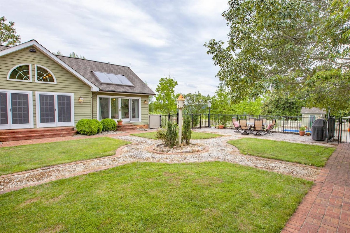 A landscaped outdoor area features a central garden path surrounded by manicured grass. The house has large windows allowing natural light, with a fenced patio in the background equipped with lounge chairs. A grill is visible, creating an inviting space for outdoor gatherings.