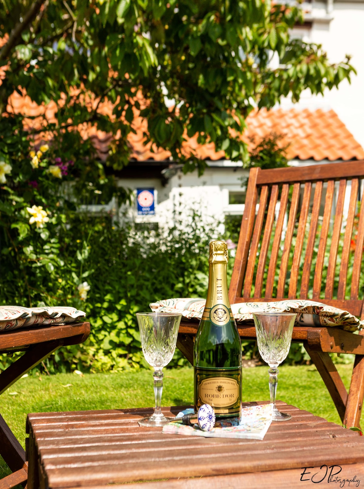 A small wooden table is set with a bottle of champagne and two crystal glasses. The table is placed on a green lawn, surrounded by wooden chairs adorned with floral cushions. The background features a cottage with a brick roof, partially obscured by lush greenery.
