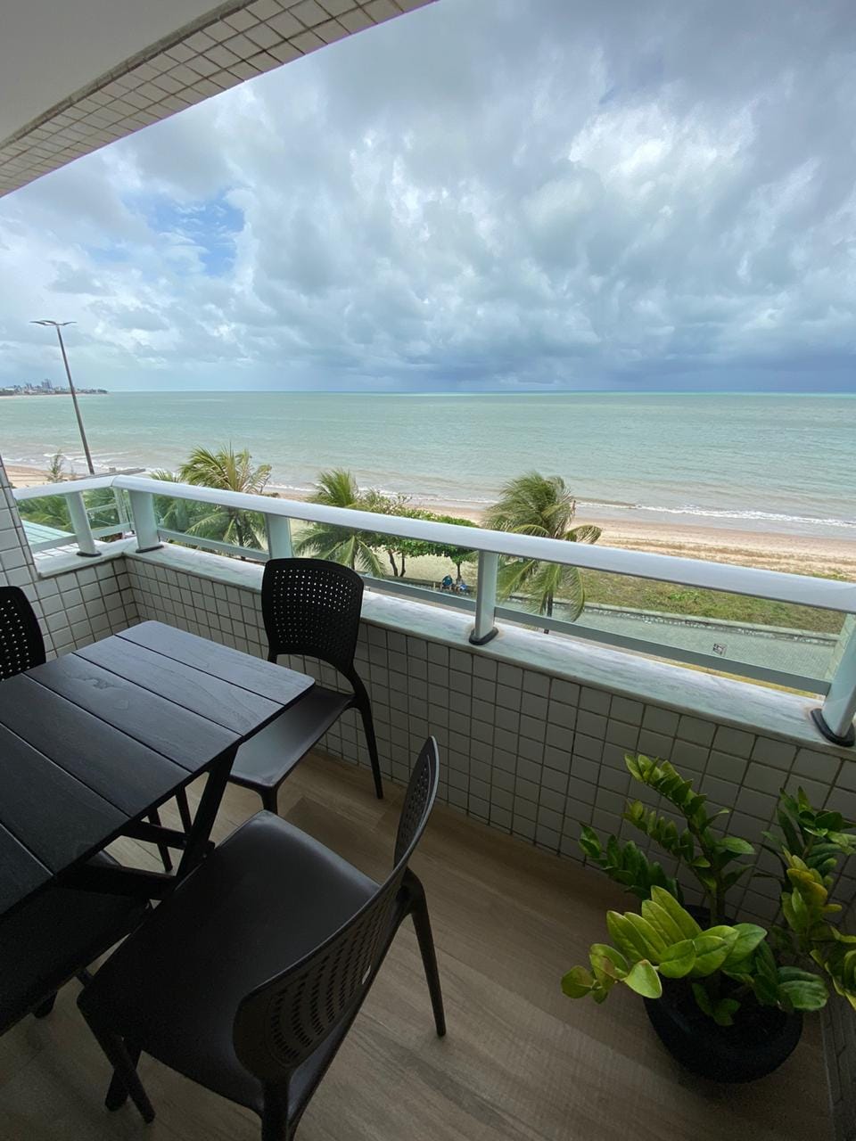 A balcony setting is presented, featuring a black table with chairs overlooking a beach. Lush greenery is seen nearby, with the coastline and water stretching out in the distance. The sky is filled with clouds, adding depth to the coastal view.