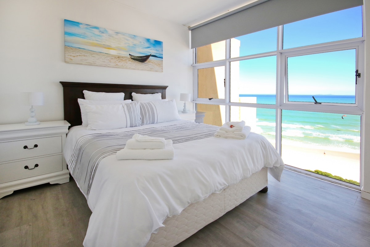 A spacious bedroom features a queen-sized bed with crisp white linens and a decorative blanket. Large windows offer ocean views, allowing natural light to fill the room. A beach-themed painting decorates the wall, and bedside tables with lamps flank the bed.