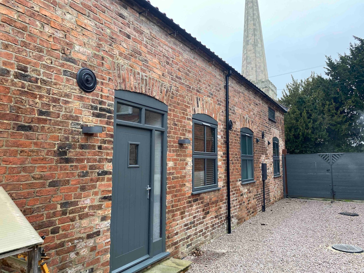 The exterior of a converted barn is presented, featuring rustic red brick walls and modern gray windows. A simple door leads into the building, with a gravel pathway in the foreground. A tall structure can be seen in the background, contributing to the urban landscape.