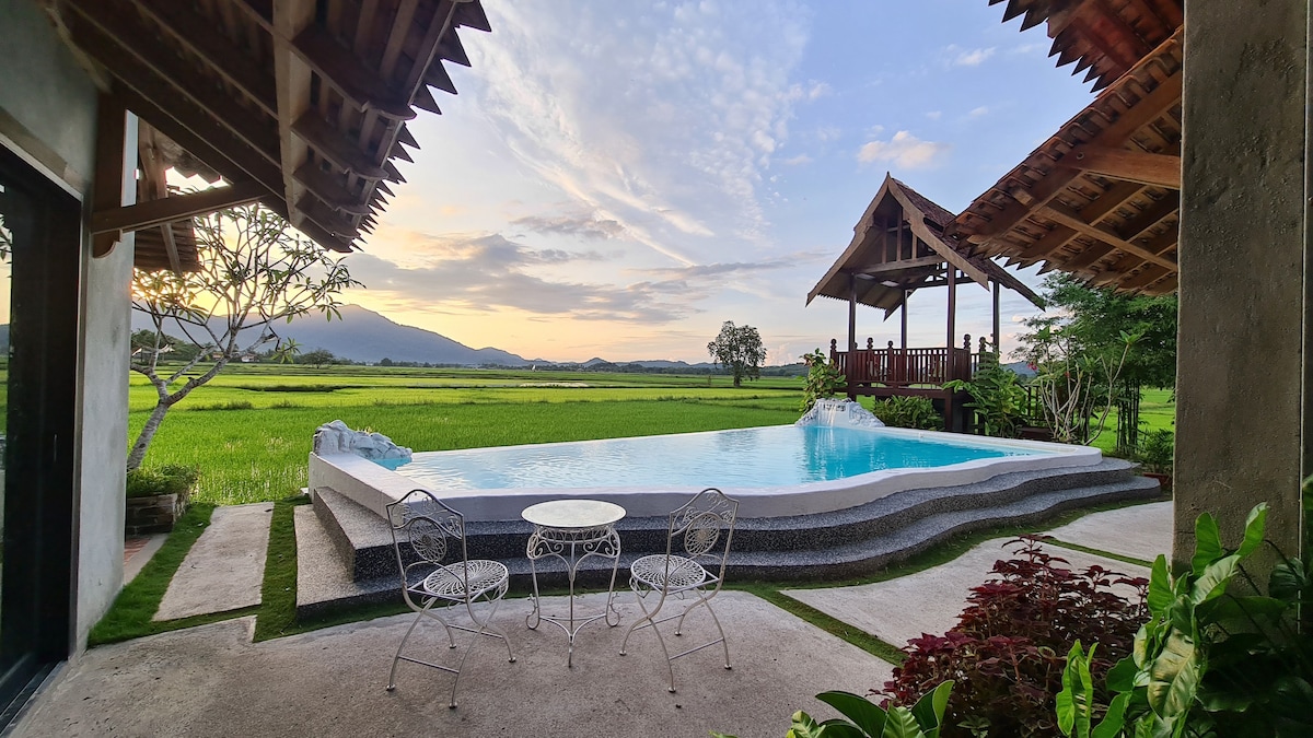 An outdoor area showcases a private infinity pool with calm blue water, framed by a stone edge. Metal patio chairs and a small table are positioned nearby, overlooking the lush paddy fields and distant mountains under a colorful sky.