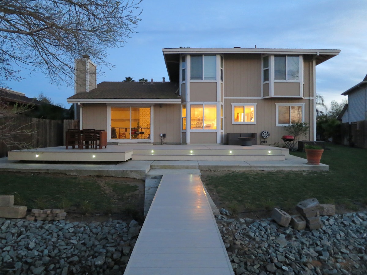 The lake house exterior is captured during twilight, showcasing a spacious deck illuminated by soft lights. A pathway leads to the water, with a landscaped yard featuring a mix of stones and grass. Large windows reflect warm interior lighting, enhancing the inviting atmosphere.
