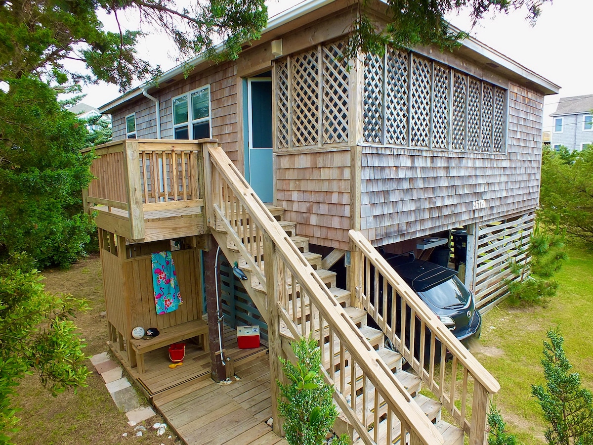 The cottage features a wooden exterior with a distinctive shingle design. A set of stairs leads to the entrance, while a shaded parking area is visible below. Lush greenery surrounds the structure, enhancing the tranquil atmosphere of the property.