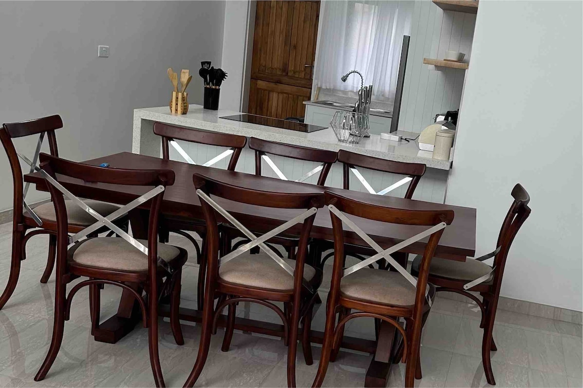 A dining area is showcased with a large wooden table surrounded by eight chairs, featuring cross-back designs. Light-colored cushions provide comfort. The kitchen space, visible in the background, includes modern appliances and a wooden door that offers a rustic touch.