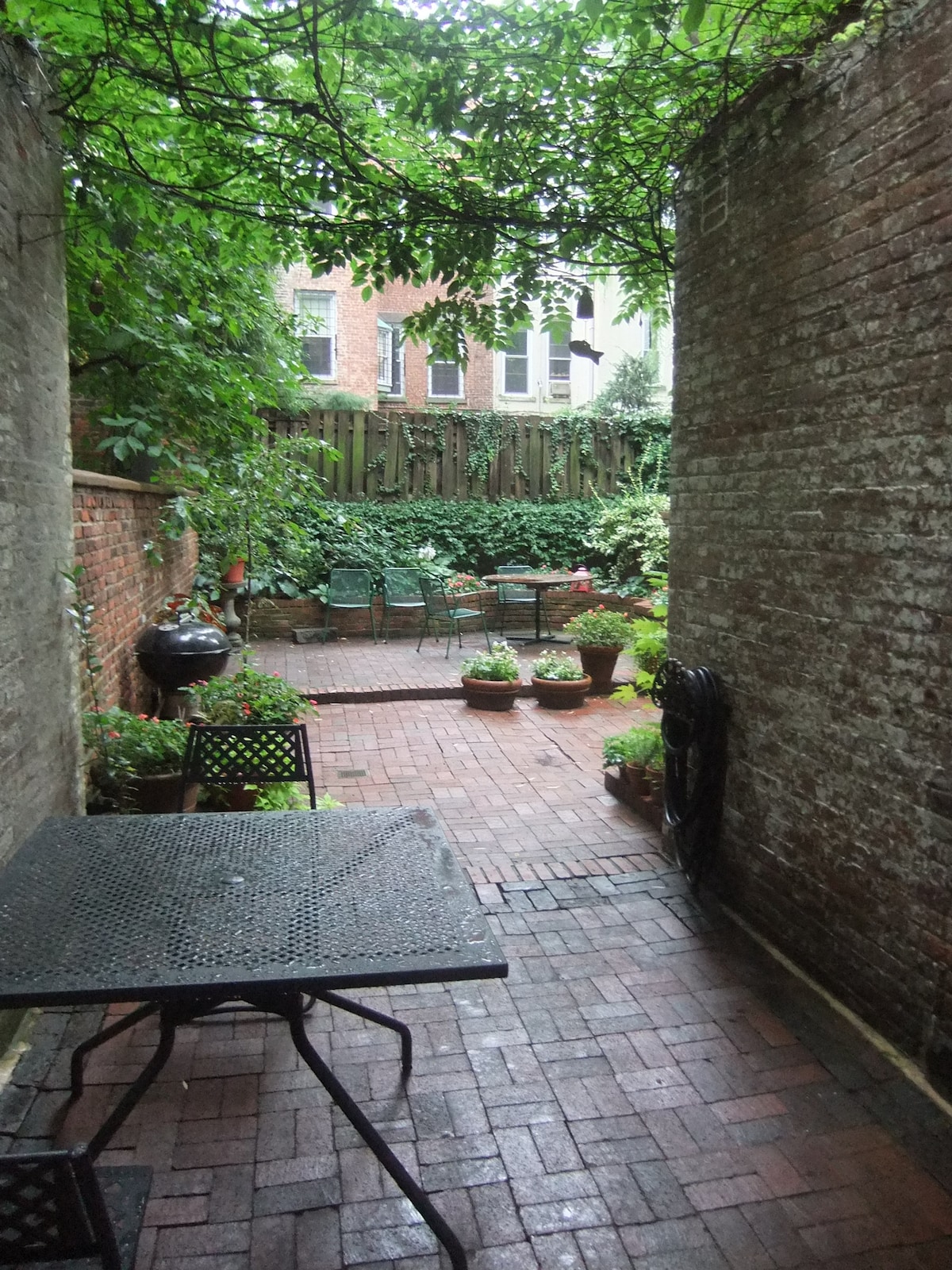 A private garden space is depicted, featuring brick pathways and potted plants. Comfortable seating is arranged amidst lush greenery and brick walls, providing a serene outdoor retreat. The area is shaded by overhanging foliage, creating a tranquil environment.