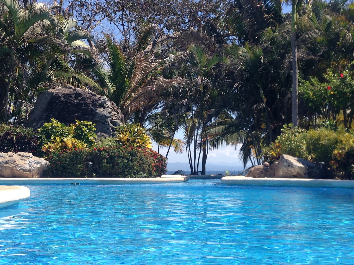 A clear blue swimming pool is surrounded by lush tropical gardens and palm trees. Brightly colored flowers and large rocks are visible along the water's edge, providing a natural and serene backdrop. The ocean can be seen in the distance, enhancing the tranquil atmosphere.