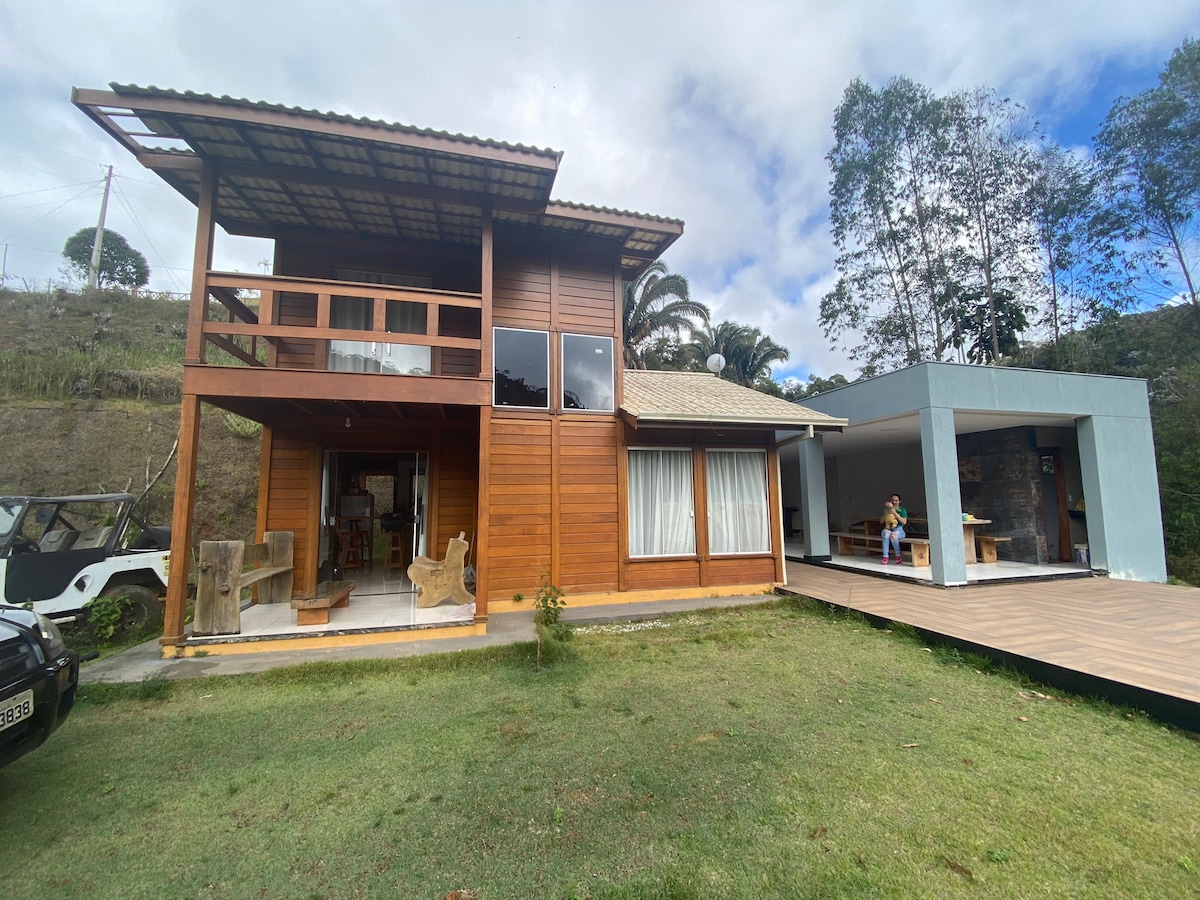 A wooden house with a sloped roof is set amidst greenery, showcasing a spacious deck and large windows. The structure features an outdoor seating area and is complemented by a well-maintained lawn, inviting relaxation in a serene mountain environment.