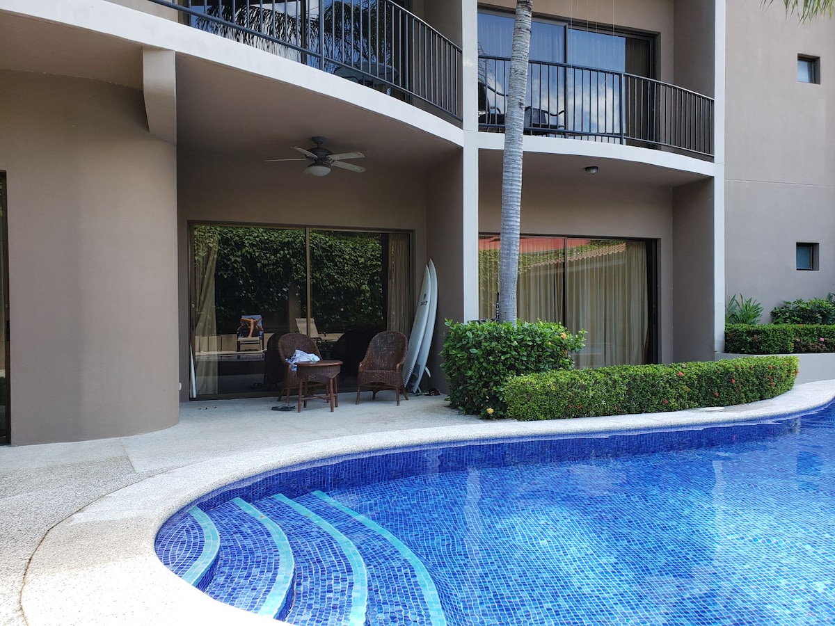 A serene outdoor space is visible, featuring a private patio with two woven chairs and a ceiling fan. A sparkling blue pool reflects the sunlight, surrounded by lush greenery. The sliding glass doors of the condo offer a view into the living area.