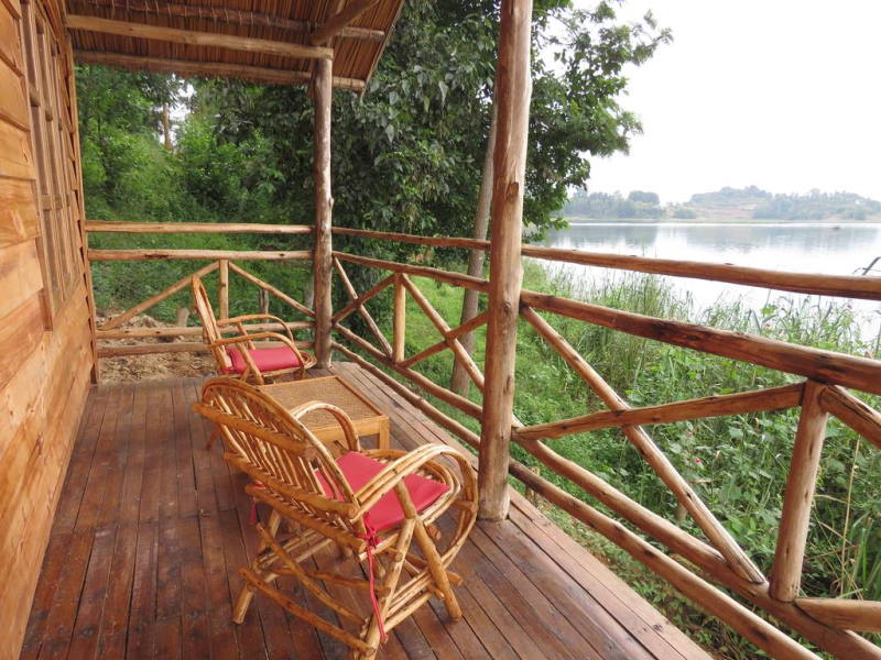 A wooden terrace is seen, furnished with two wicker armchairs and a small table, all featuring red cushions. The surrounding area is lush with greenery, offering a view of the calm lake just beyond the railing.