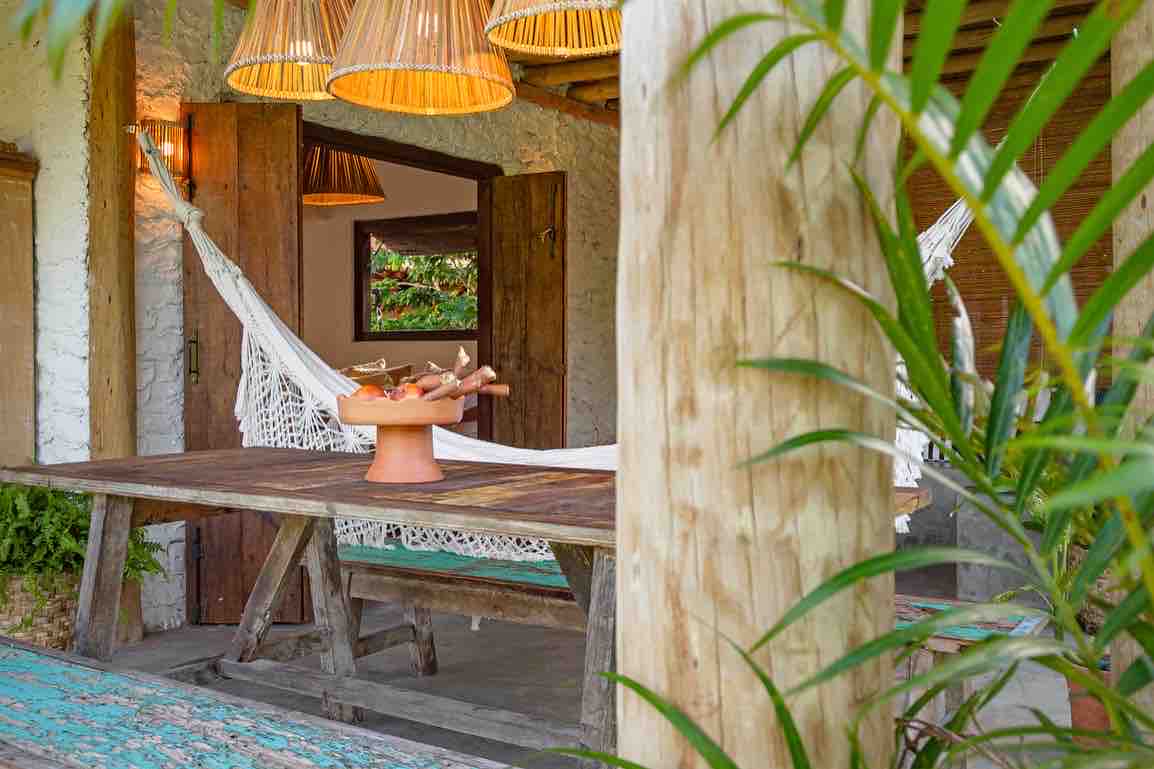 A rustic dining table made of wood is positioned under warm lighting from woven pendant lamps. A hammock is stretched nearby, enhancing the tranquil setting. Lush green plants frame the scene, providing a natural feel.