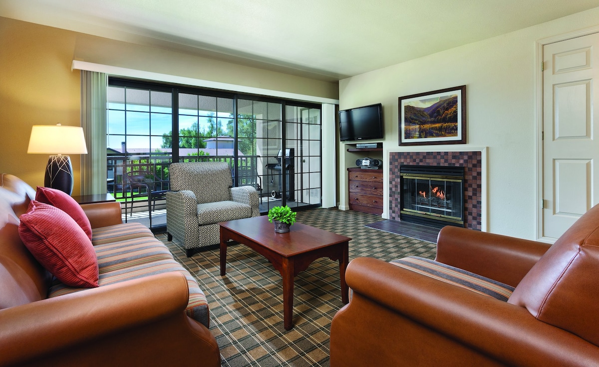 A living area features comfortable seating with two armchairs and a sofa, all arranged around a wooden coffee table. A gas fireplace provides warmth, while sliding glass doors offer natural light and access to a balcony. A television is mounted on the wall opposite the seating.