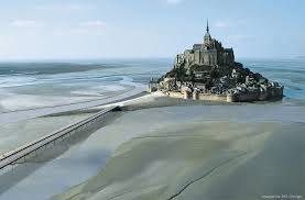 A view of Mont Saint Michel is presented, showcasing its historic architecture rising from a vast tidal landscape. The surrounding waters reflect shades of blue and gray, with a narrow causeway leading towards the island. The sky is clear, enhancing the serene atmosphere of the scene.