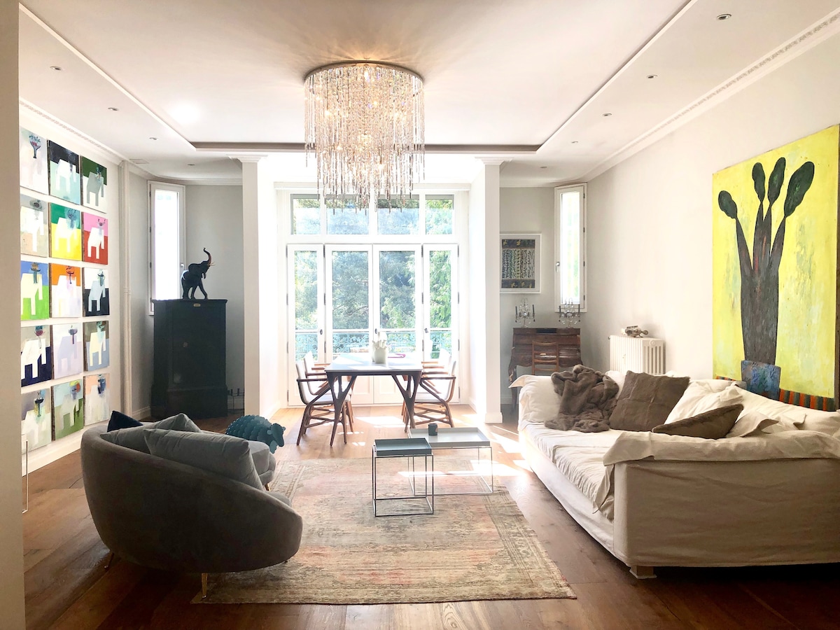 A spacious living area is adorned with contemporary artwork, featuring a large canvas showcasing a tree. Oversized seating options, including a plush armchair and a sectional sofa, provide comfort. A glass coffee table sits on a textured rug, while natural light filters through large windows with scenic views.