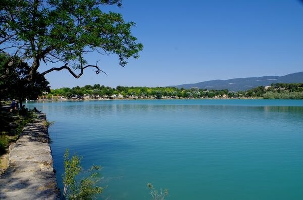 A serene view of a calm lake framed by lush greenery. The water's surface shimmers in various shades of blue, reflecting the clear sky. In the distance, trees line the shore, adding a sense of tranquility to the landscape.