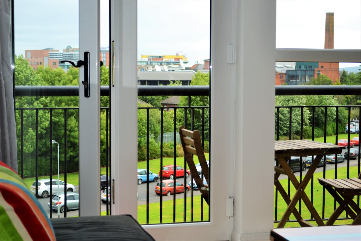 A view from a bright living space displays a private balcony with a bistro table and chairs. Beyond, city buildings and greenery create a scenic backdrop. Various colorful cars are visible parked along a winding driveway under a cloudy sky.