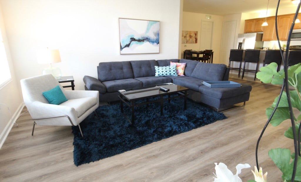 A comfortable living area is presented, featuring a modern sectional sofa and a stylish armchair. A dark coffee table is positioned atop a plush blue area rug. Light walls and a piece of wall art enhance the ambience, with a glimpse of the open kitchen and dining area in the background.