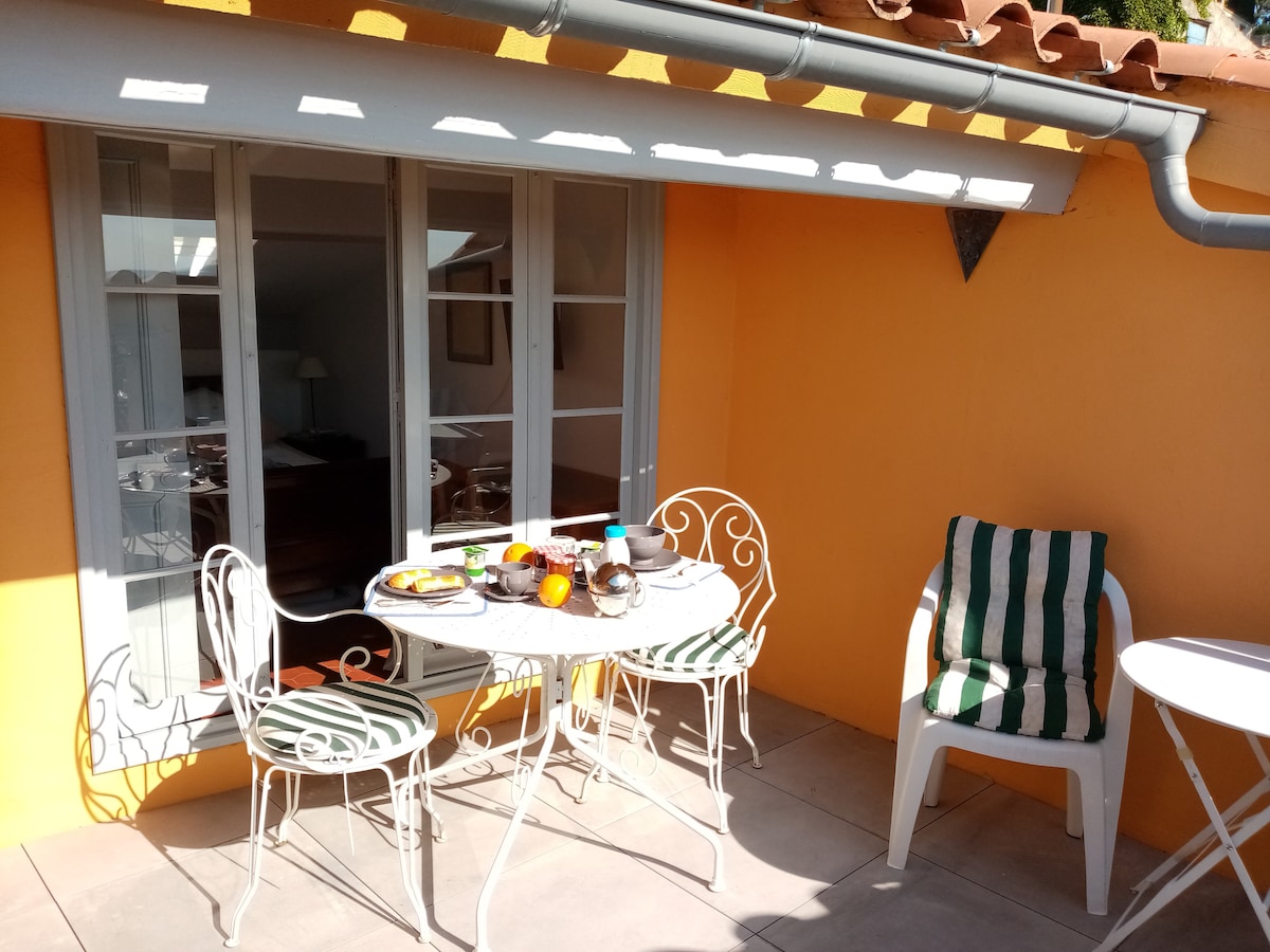 A charming terrace is featured, furnished with a round table and four metal chairs. Bright orange walls provide a vibrant backdrop, while a striped lounge chair adds comfort. Partial sunlight illuminates the space, creating a welcoming area for outdoor dining or relaxation.