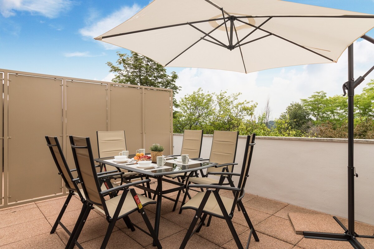 An outdoor dining area is furnished with a large table surrounded by six folding chairs. A large umbrella provides shade for the seating area. Lush greenery is seen in the background, enhancing the peaceful outdoor setting.