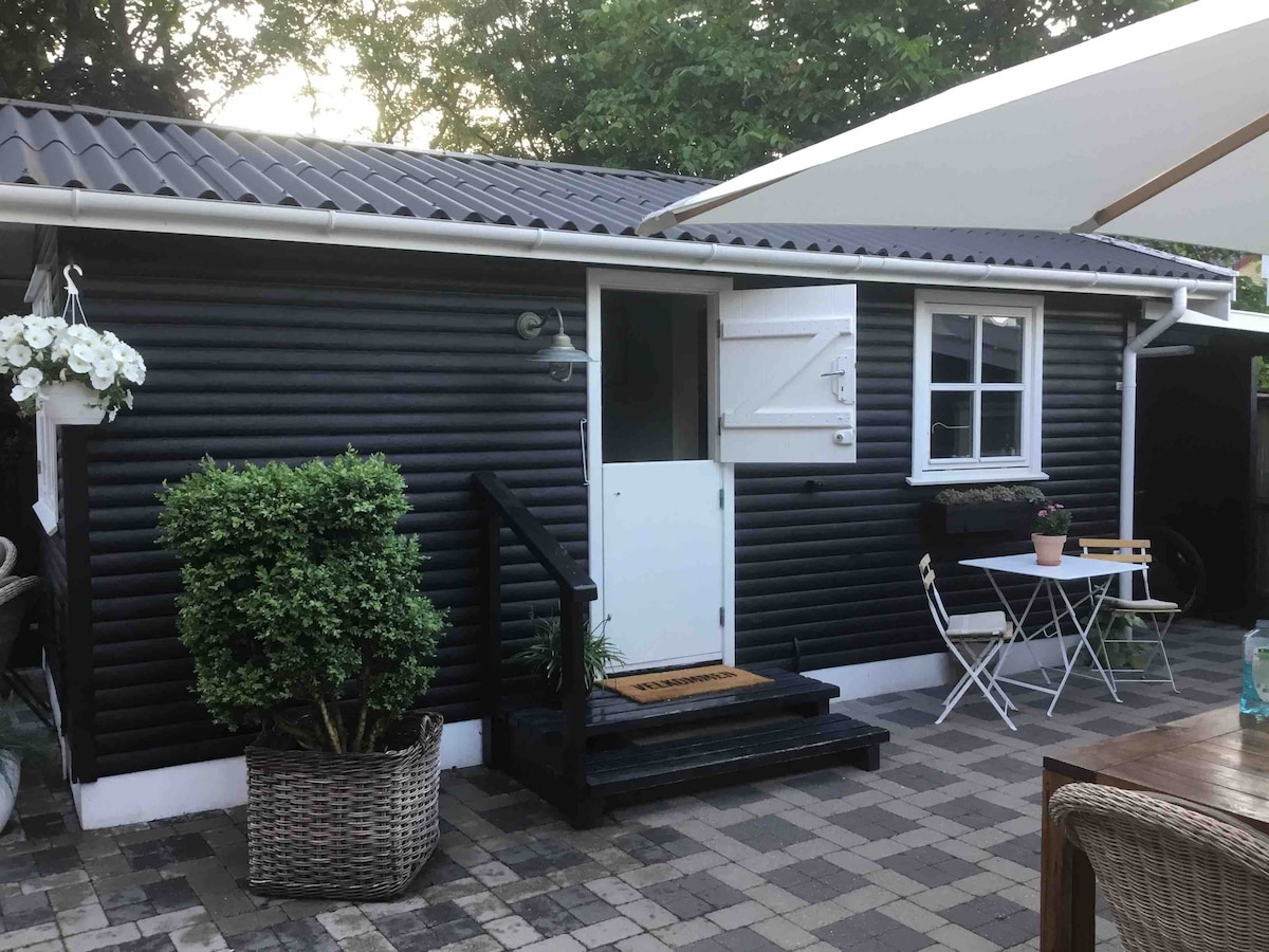 A charming black exterior cottage is depicted, featuring a white door and a small window. A staircase leads to the entrance, while a potted plant adorns the side. A small outdoor table and chairs are arranged in front, with foliage and a shaded area suggested by the nearby awning.