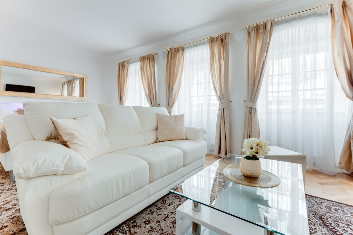 A spacious living area features a light-colored sofa adorned with pillows, complemented by a glass coffee table. Large windows adorned with soft drapes allow natural light to fill the room, creating an airy and bright atmosphere.