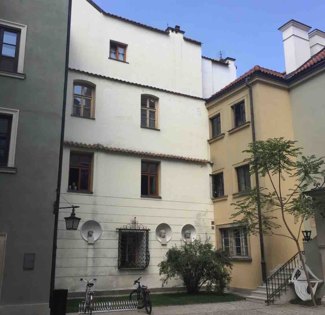 The image shows a quiet courtyard surrounded by three buildings with varying architectural styles. Windows are visible, some with dark frames. A bike is parked near a decorative fence. Green foliage adds a touch of nature to the serene scene.