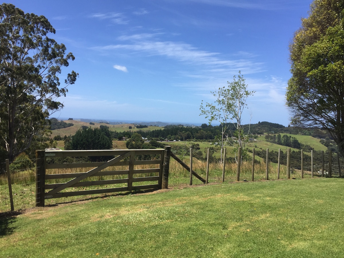 A grassy area features a wooden fence leading towards a gate, providing views of the rolling hills and distant sea. Lush greenery surrounds the landscape, with trees partially framing the scene and a clear blue sky enhancing the serene rural setting.