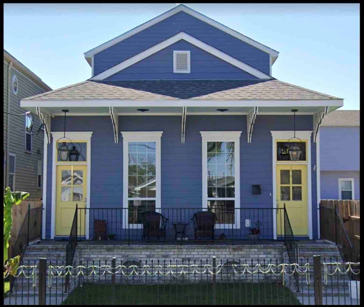 A two-unit residence features a blue exterior with a symmetrical design. Yellow doors provide a bright contrast to the siding. The front porch is furnished with several chairs, and a well-maintained lawn is visible in front of the property.