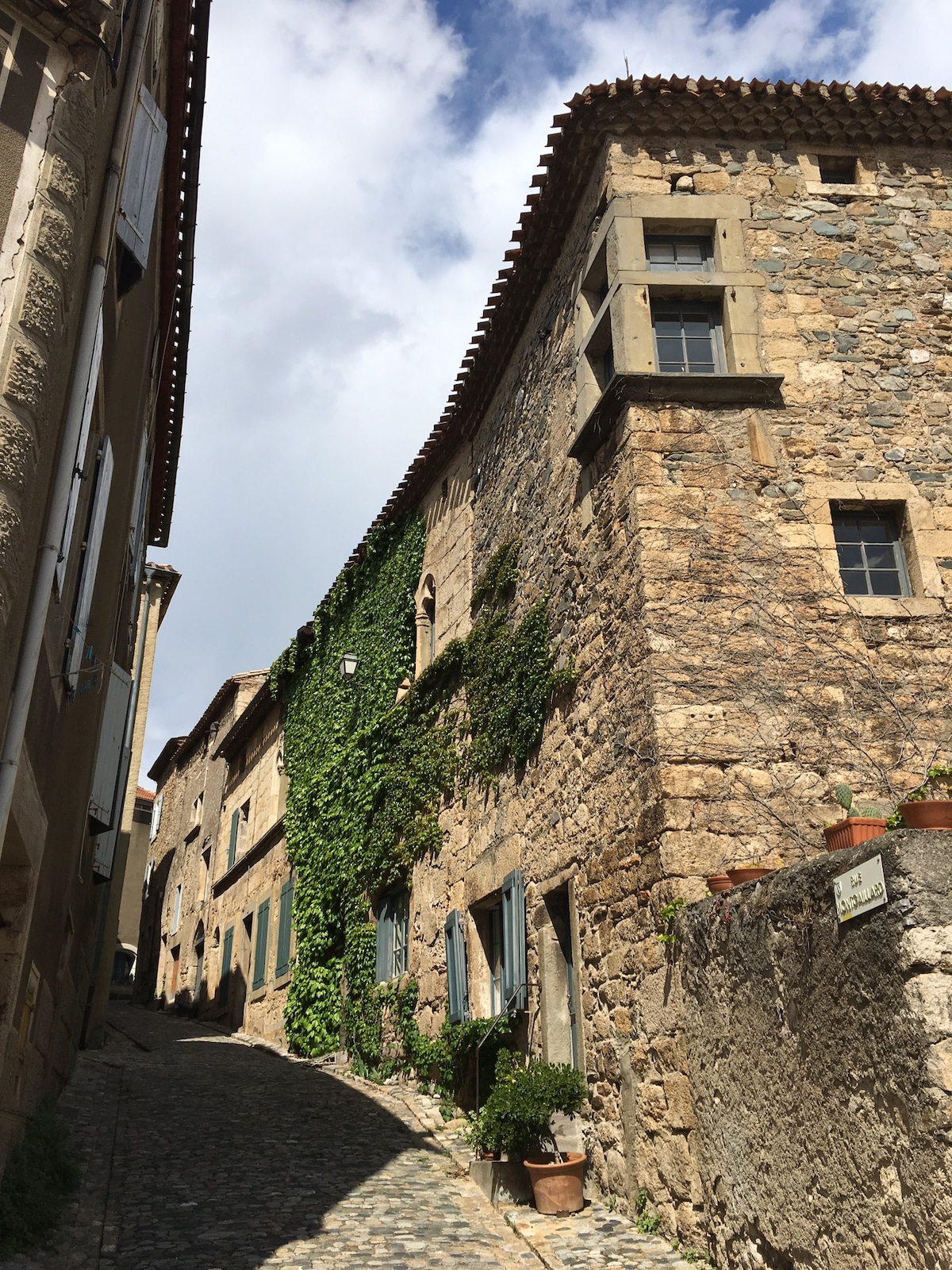 An ancient stone building is seen, characterized by ivy climbing its textured facade. Windows are framed with blue shutters, providing a rustic charm. A narrow, cobblestone street leads towards the structure, flanked by additional historic architecture, under a partly cloudy sky.