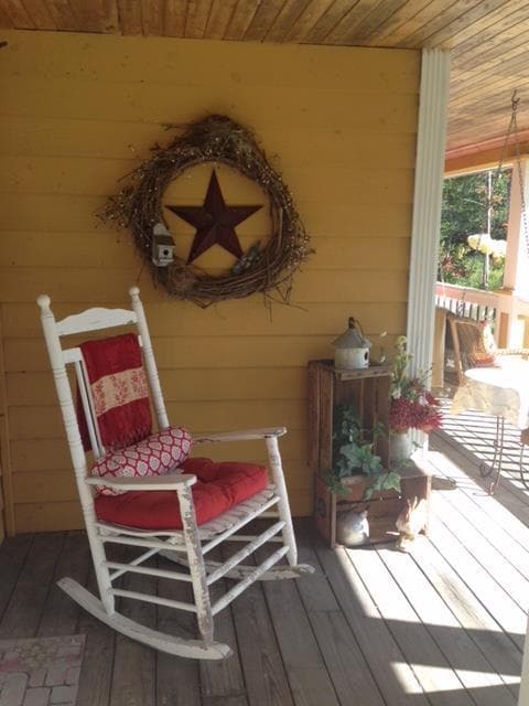 A rocking chair with a red cushion is positioned on a wooden porch, accompanied by a decorative wreath featuring a star. A small wooden crate holds potted plants, enhancing the welcoming ambiance. Sunlight streams in, illuminating the wooden flooring and surrounding elements.