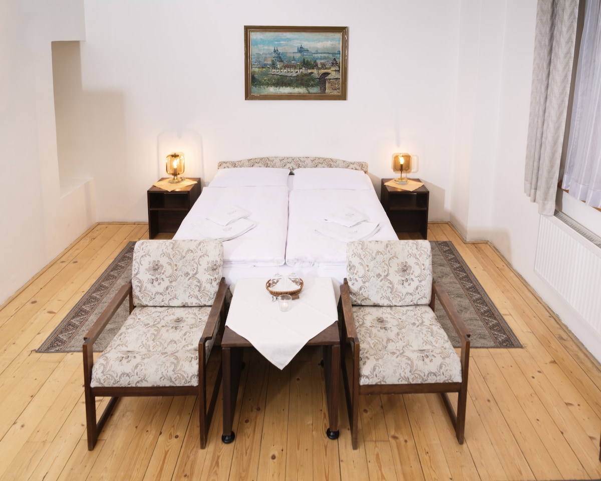 A spacious sleeping area features a large bed dressed in fresh linens. Two patterned armchairs flank a small table in front of the bed. A framed artwork adorns the wall, and light filters through a nearby window, highlighting the wooden floor.