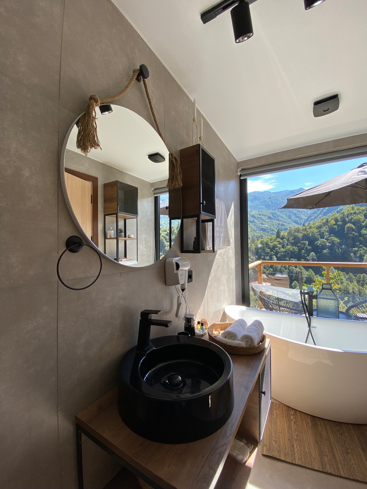 A modern bathroom features a round mirror with natural accents above a sleek black sink. A wooden vanity holds towels and amenities. Natural light enters through a large window, offering a view of the surrounding mountains and a balcony with a shaded area.