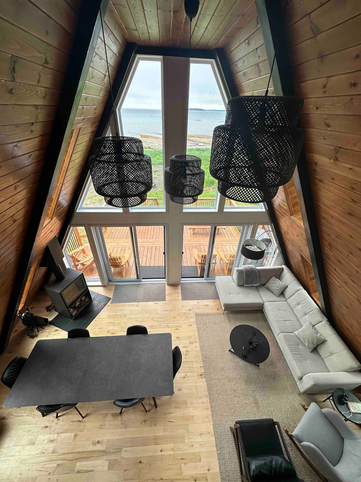 An open living space features tall A-frame ceilings with contemporary hanging light fixtures. Large glass doors offer views of the sea and lead to an outdoor deck. A modern sofa faces a coffee table, while a dining table for six is positioned nearby on warm wooden flooring.