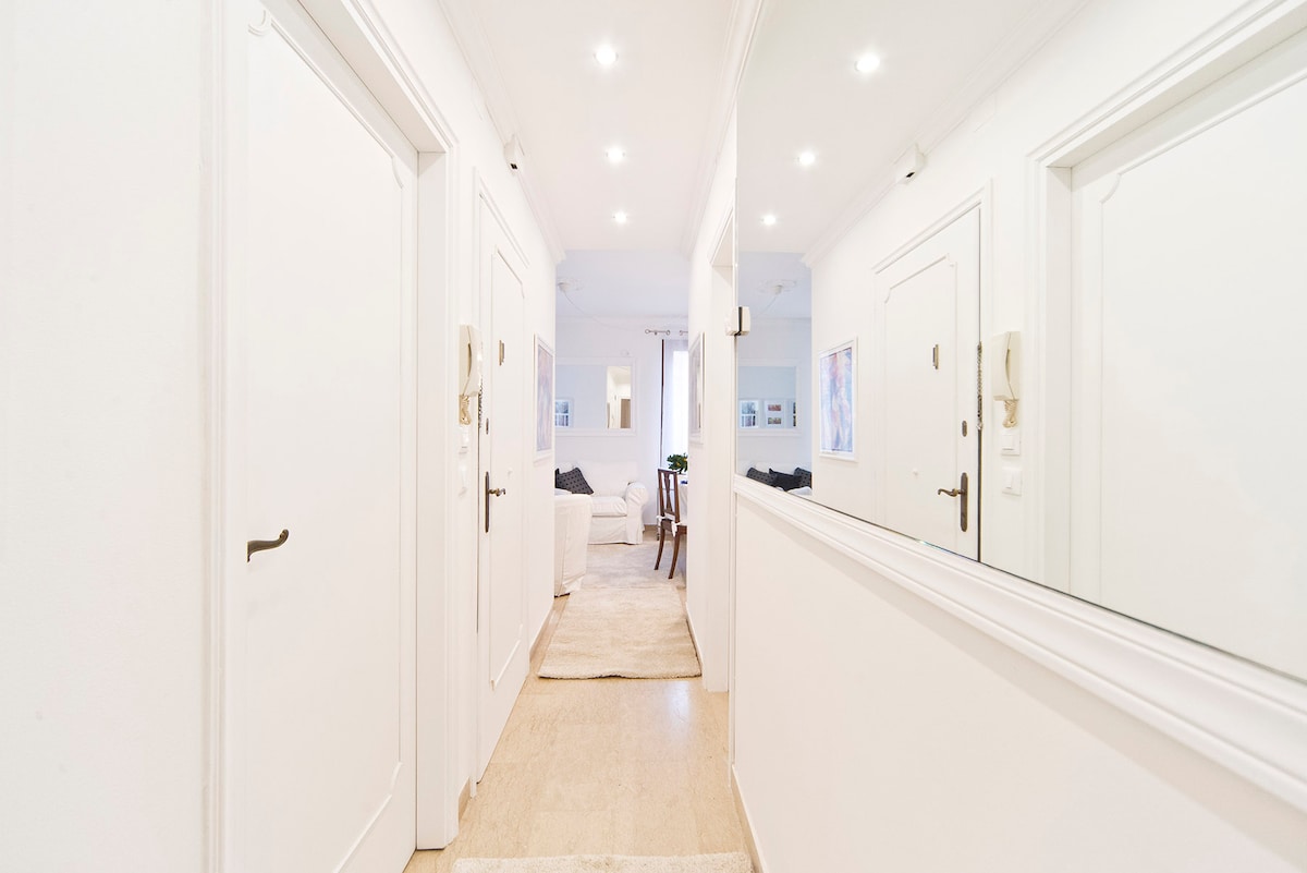 A well-lit hallway is shown, featuring white walls and light wood flooring. A large mirror reflects the space, and doorways to rooms are visible on both sides. Soft lighting fixtures enhance the brightness, contributing to an open and inviting atmosphere.