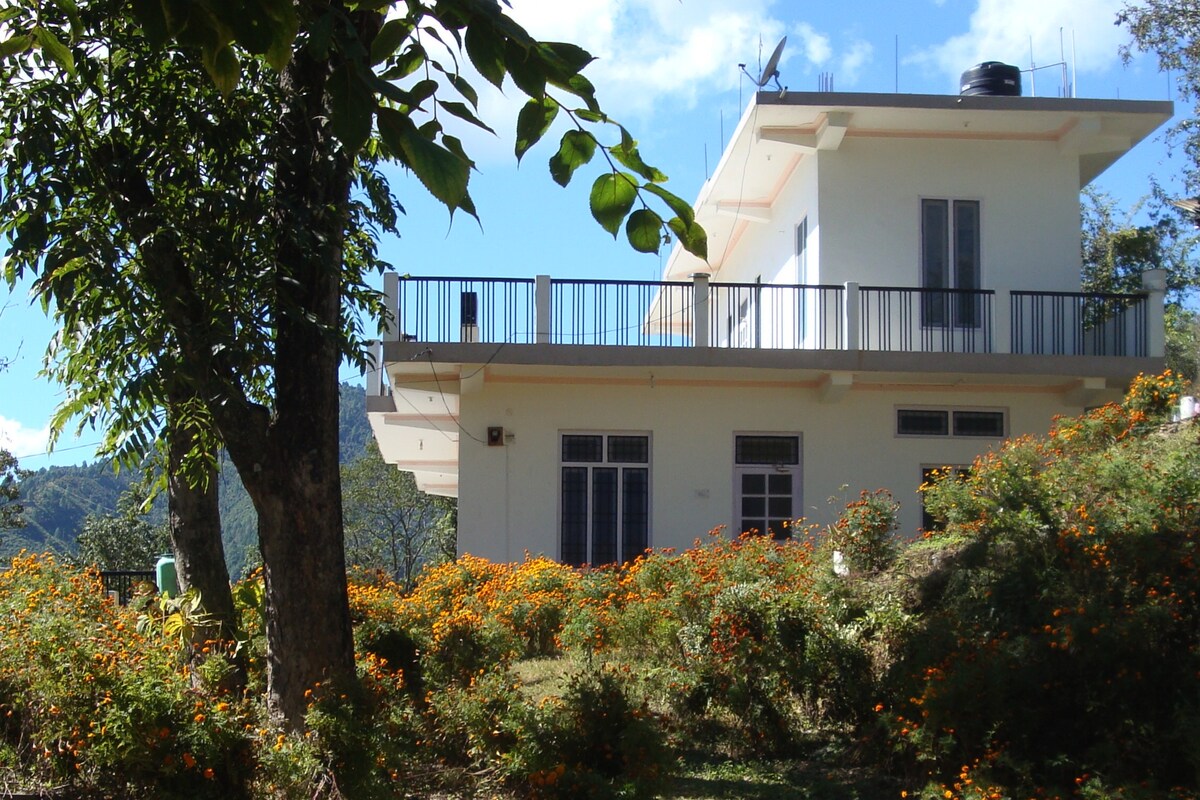 A modern two-story home is surrounded by lush greenery and vibrant flowering plants. A spacious balcony extends from the upper level, offering views of the scenic landscape. The property is integrated with nature, providing a serene environment for relaxation.