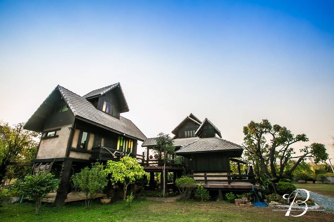 Two traditional Thai houses are situated on elevated wooden platforms, surrounded by lush greenery. The larger house features multiple levels and a gabled roof, while the smaller house displays a simple design. A serene sky complements the tranquil setting.