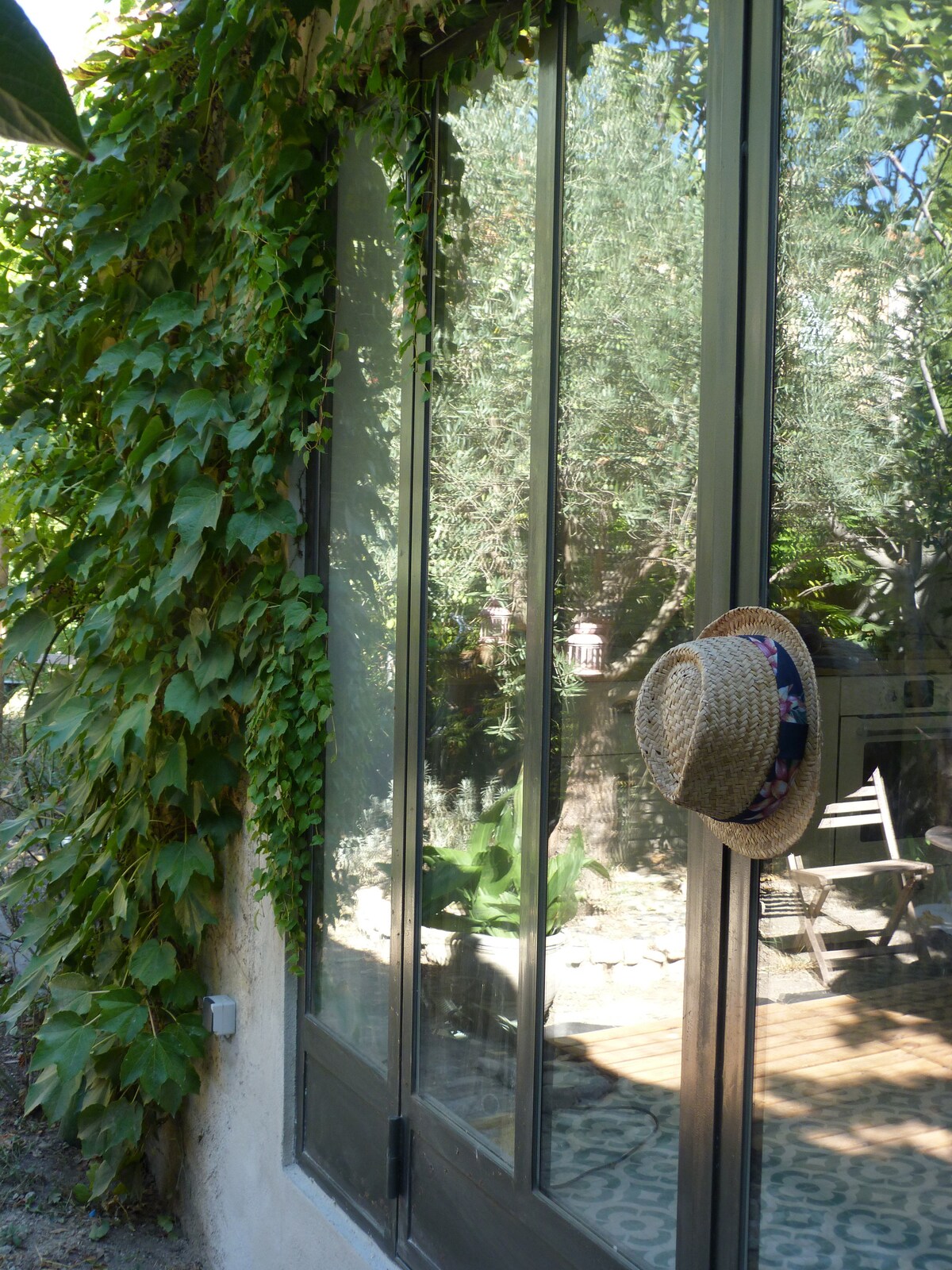 A glass door framed by lush green vines leads into a serene space. A straw hat hangs casually on the door, suggesting a relaxed atmosphere. In the background, sunlit patterns hint at a tiled floor and outdoor greenery.