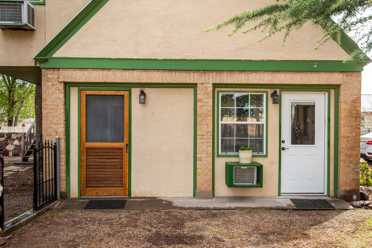 The exterior of a charming two-story building is presented, featuring a green-accented facade. A front door with a wooden screen stands to the left, while a white door is visible on the right. A small window air conditioning unit is installed beneath a window with a simple planter.