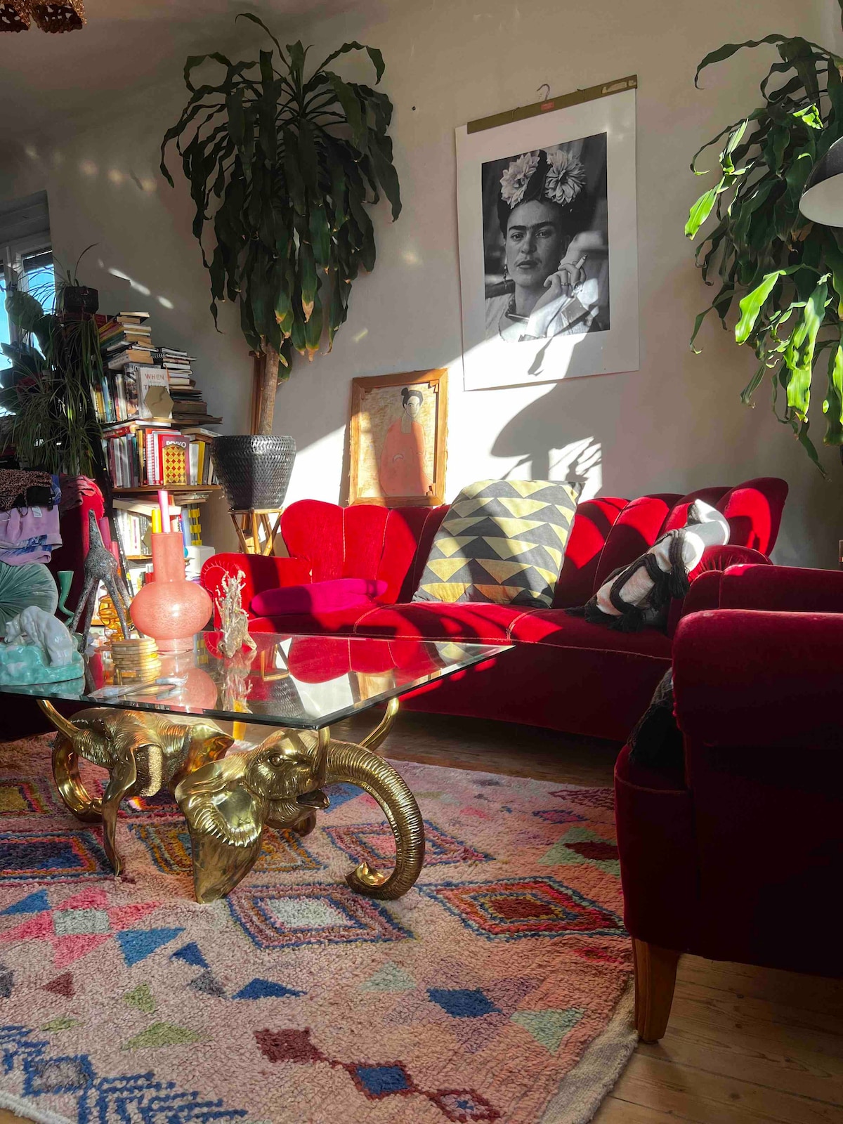 A vibrant living area features a plush red sofa, accented with decorative pillows, alongside a glass coffee table with a sculptural base. Lush plants provide greenery, while a colorful rug adds warmth to the wooden floor. Natural light filters through the space, creating a welcoming ambiance.