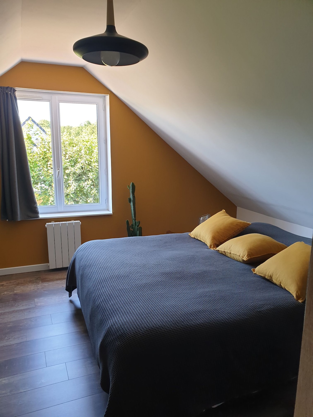 A spacious bedroom features a sloped ceiling and warm orange walls. A comfortable bed is dressed with dark bedding and complemented by two yellow pillows. A window allows natural light to illuminate the room, while a decorative plant provides a touch of greenery.