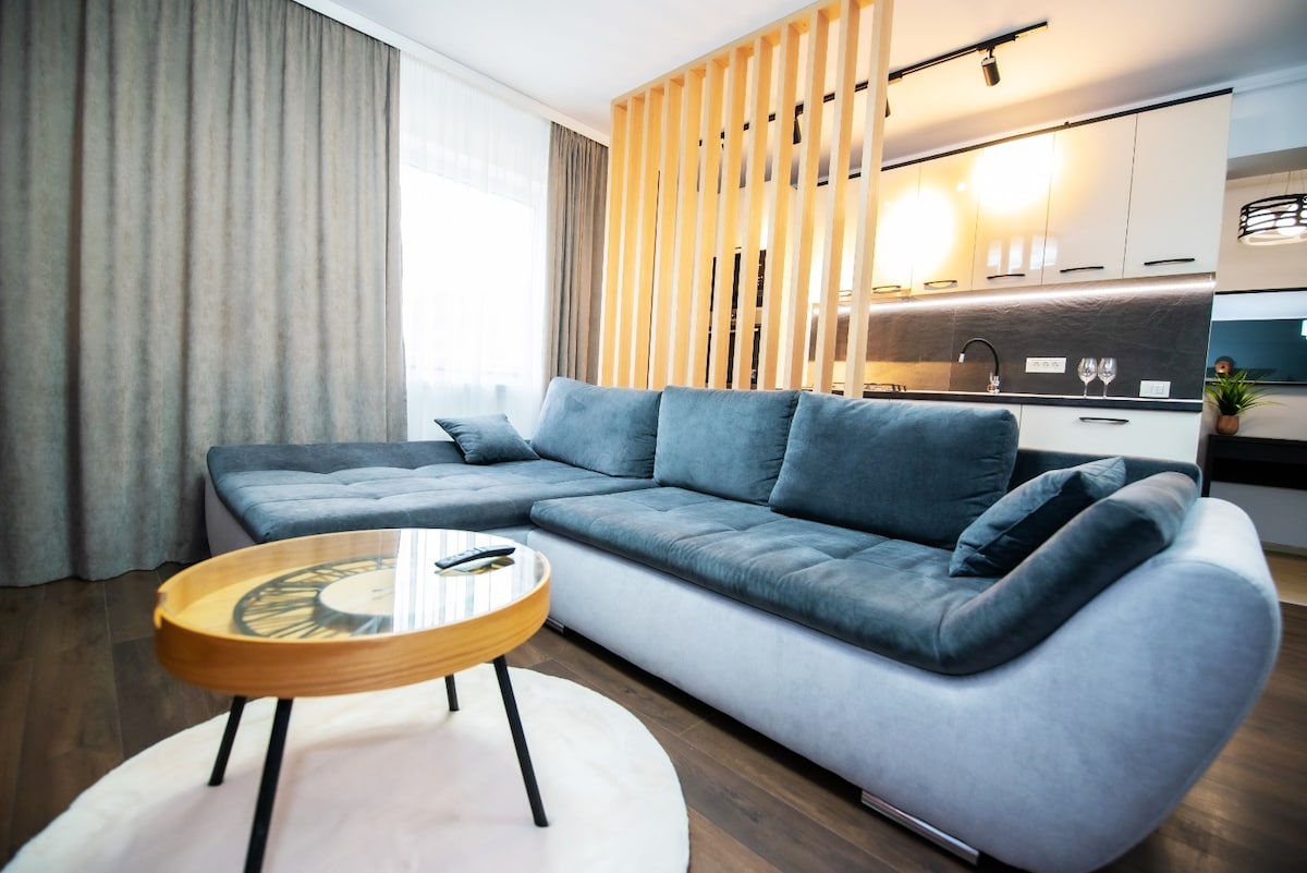 A spacious living area features a large, grey sectional couch positioned against a wooden partition. A round coffee table with a wooden top sits on a soft area rug. Natural light is invited through sheer curtains, enhancing the inviting ambiance of the room.