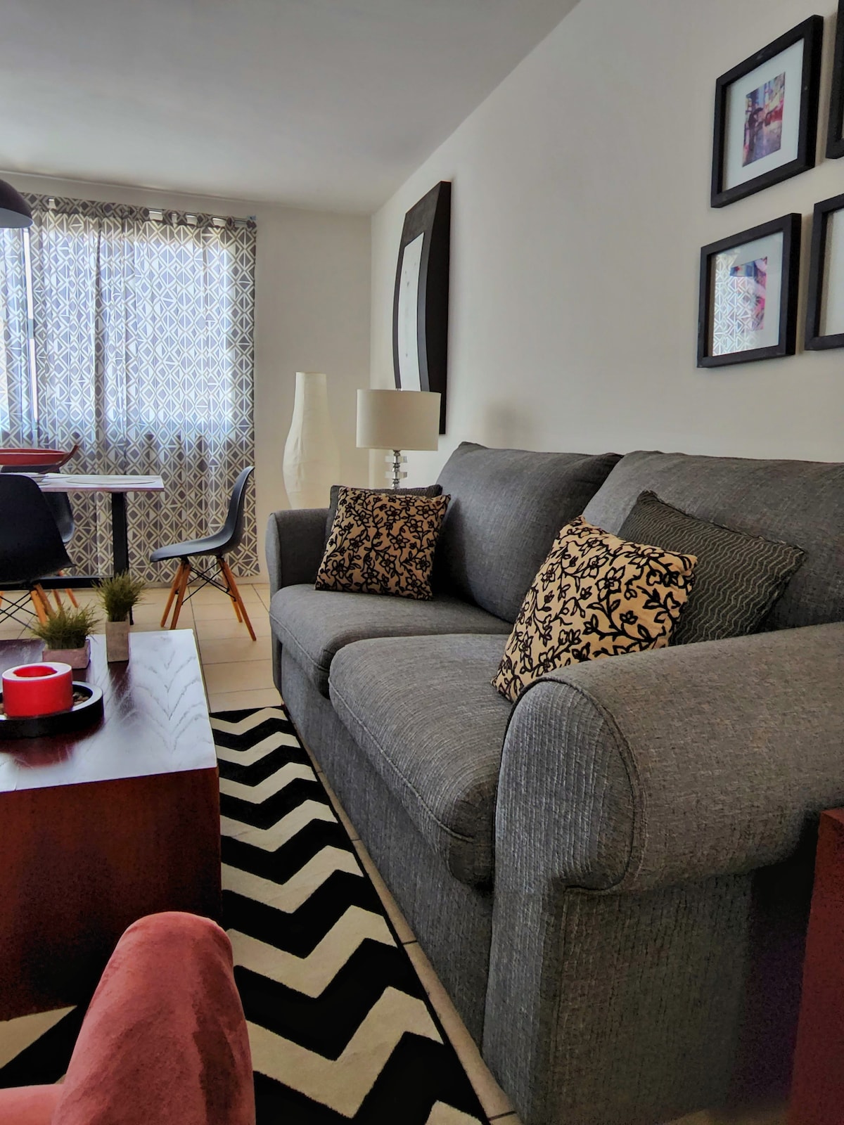 A cozy living area features a comfortable gray sofa adorned with patterned pillows. A wooden table sits nearby, complemented by black dining chairs and bright window treatments. A geometric rug adds texture, while decorative items enhance the contemporary decor.