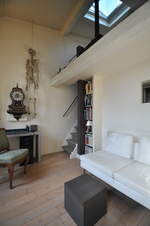 The interior features high ceilings with a skylight, illuminating a minimalist design. A white sofa is positioned beside a compact coffee table, and a bookshelf displays various books. A decorative skeleton hangs on the wall, adding a unique element to the cozy space.